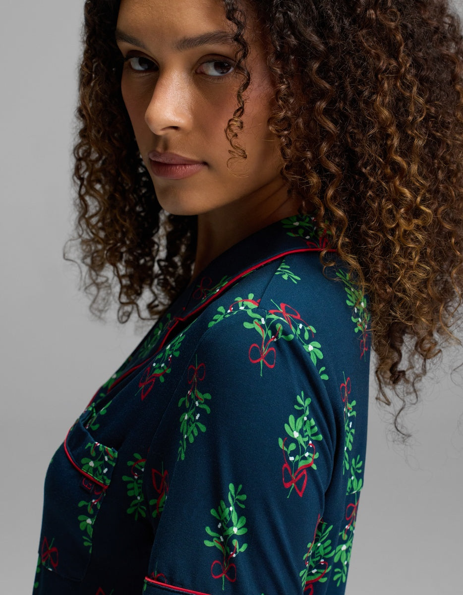 A person with curly hair wearing a dark blue Women’s Bamboo Stretch-Knit Short Sleeve Pajama Top featuring a pattern of mistletoe and red bows.