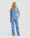 A woman with long blonde hair wears the Cozy Earth Women's Bamboo Stretch-Knit Short Sleeve & Pant Pajama Set in blue with white floral patterns. She stands barefoot against a plain light background.|Color:Garden Toile in Sailor