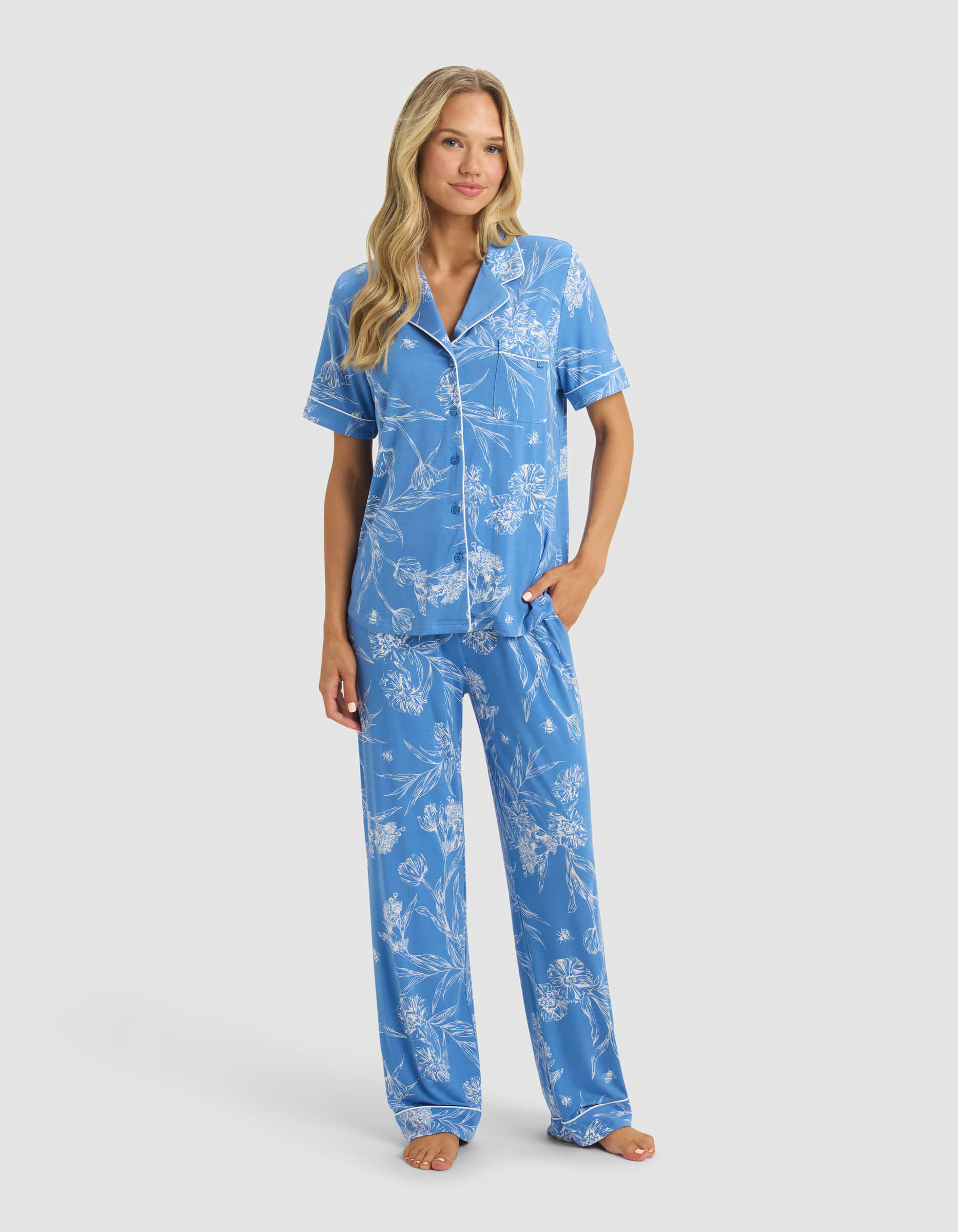 A woman with long blonde hair wears the Cozy Earth Women's Bamboo Stretch-Knit Short Sleeve & Pant Pajama Set in blue with white floral patterns. She stands barefoot against a plain light background.