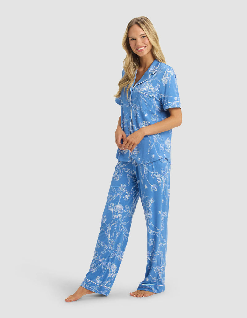 A woman with long blonde hair smiles barefoot against a plain light background, wearing Cozy Earth's Women's Bamboo Stretch-Knit Short Sleeve & Pant Pajama Set in light blue with a white floral pattern.