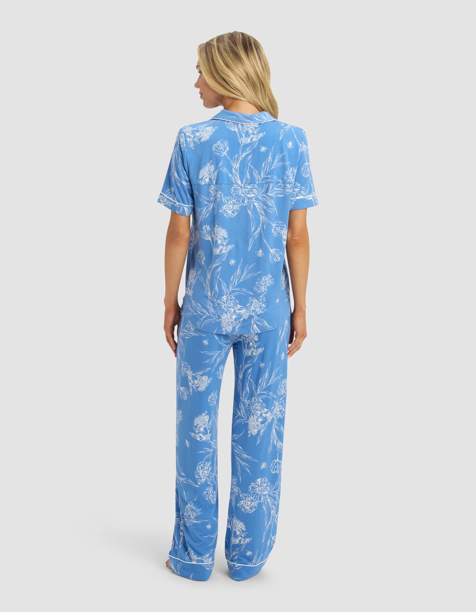 A woman with long blonde hair stands facing away against a plain light background, wearing Cozy Earth's Women's Bamboo Stretch-Knit Short Sleeve & Pant Pajama Set in blue with a white floral pattern.