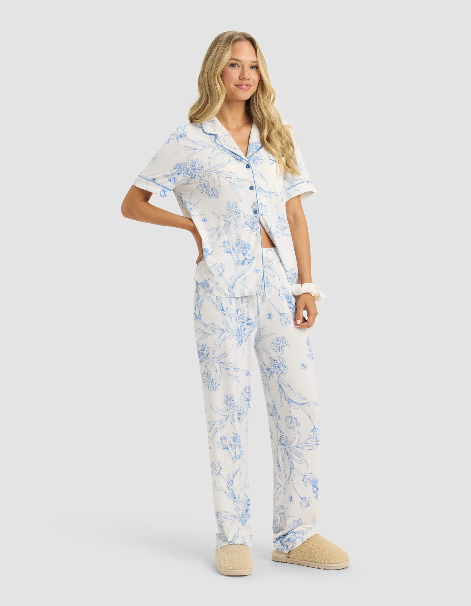 A woman with long blonde hair wears the Cozy Earth Women's Bamboo Stretch-Knit Short Sleeve & Pant Pajama Set in white with blue floral prints and beige slippers, standing casually with one hand on her hip.
