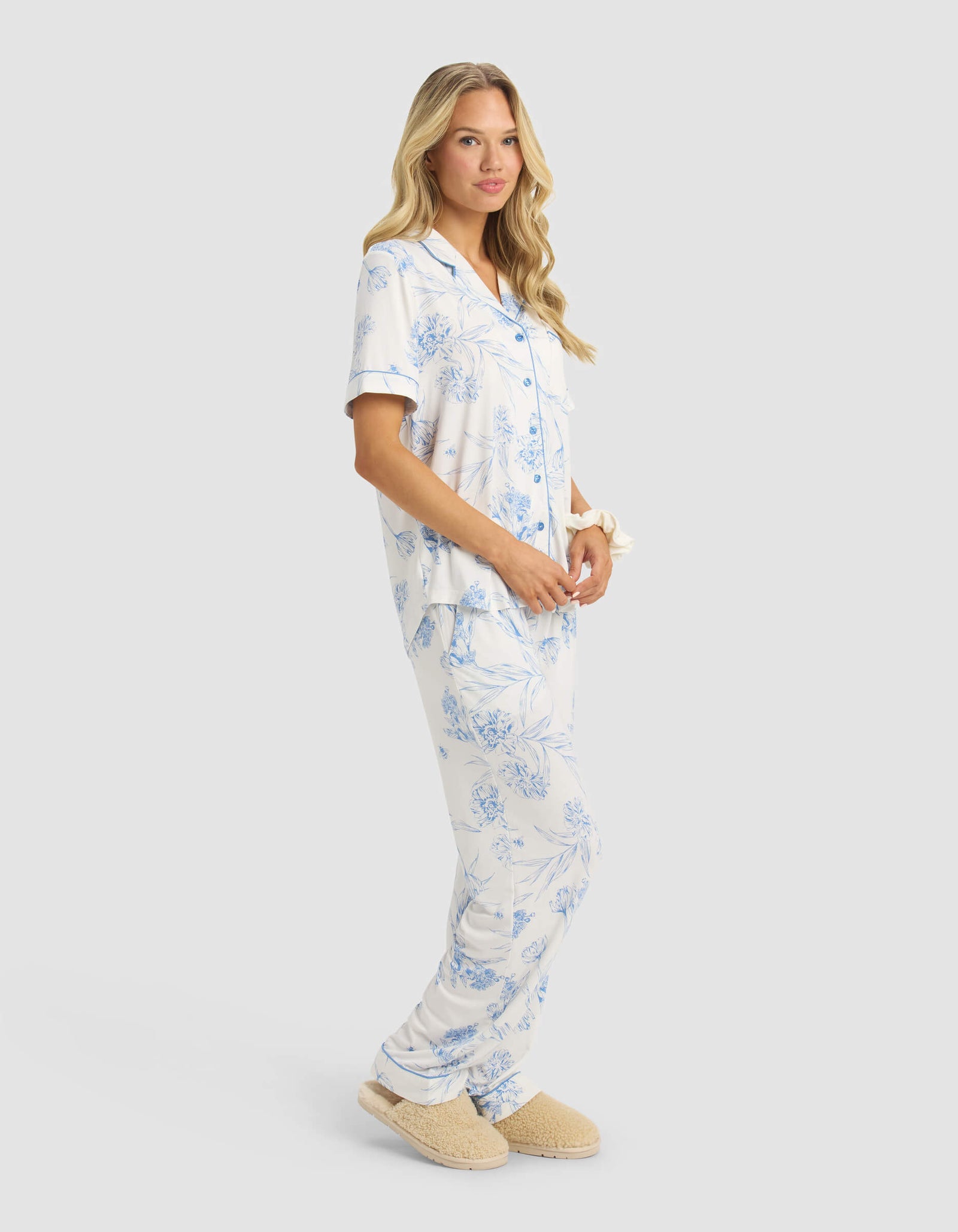A woman with long, wavy blonde hair and a relaxed expression models the Cozy Earth Women's Bamboo Stretch-Knit Short Sleeve & Pant Pajama Set in light blue and white floral against a plain light background, paired with beige slippers.