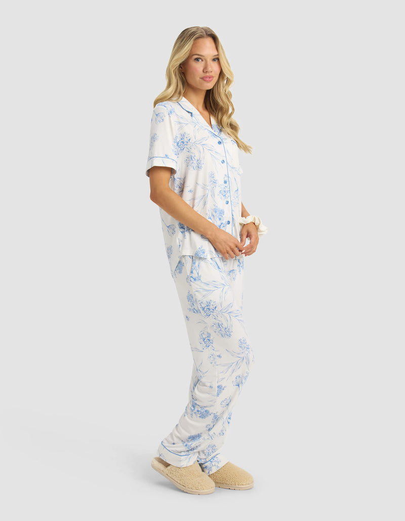 A woman with long, wavy blonde hair and a relaxed expression models the Cozy Earth Women's Bamboo Stretch-Knit Short Sleeve & Pant Pajama Set in light blue and white floral against a plain light background, paired with beige slippers.