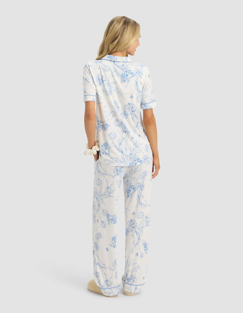 A woman with blonde hair faces away, wearing the Cozy Earth Women's Bamboo Stretch-Knit Short Sleeve & Pant Pajama Set in white with light blue floral print. She has a scrunchie on her wrist and is wearing fuzzy slippers.