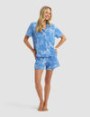 A smiling woman stands barefoot in Cozy Earth's Women's Bamboo Stretch-Knit Short Sleeve Pajama Set, featuring a light blue button-up top and matching floral shorts, against a plain light background.|Color:Garden Toile in Sailor