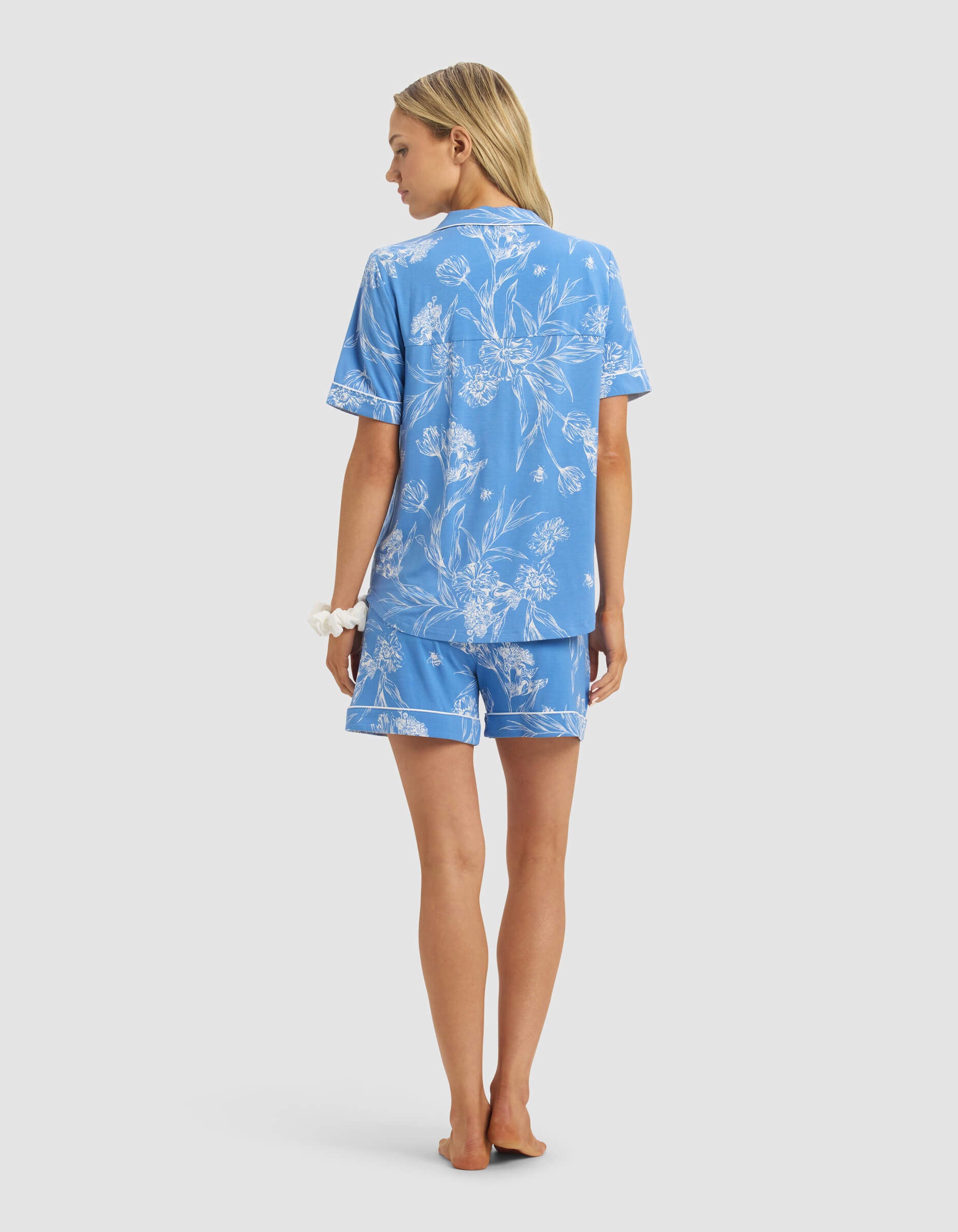 A woman with long blonde hair wears the Cozy Earth Women's Bamboo Stretch-Knit Short Sleeve Pajama Set in light blue with white floral patterns. She stands barefoot facing away, a white scrunchie on her wrist, against a plain white background.|Color:Garden Toile in Sailor