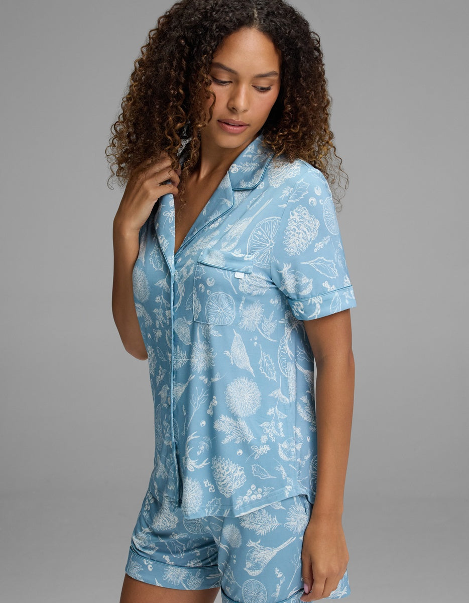 A woman wearing a Cozy Earth Women’s Bamboo Stretch-Knit Short Sleeve Pajama Top in a light blue pattern of white pinecones, citrus slices, and sprigs.