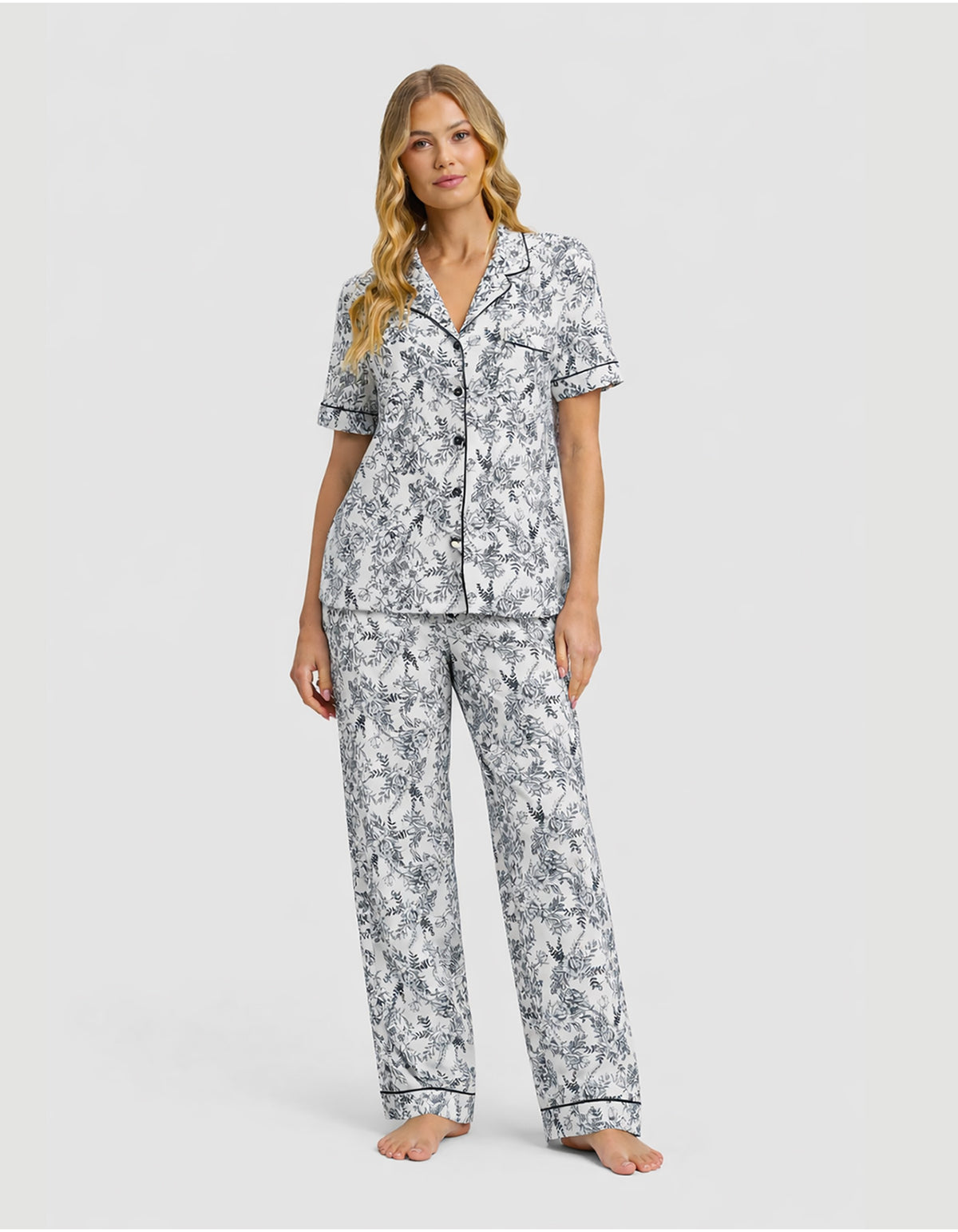 A woman stands barefoot against a plain light background, wearing the Cozy Earth Women’s Bamboo Stretch-Knit Short Sleeve & Pant Pajama Set in Jet Black Toile—a matching white pajama set with a black floral pattern and a short-sleeved button-up top.
