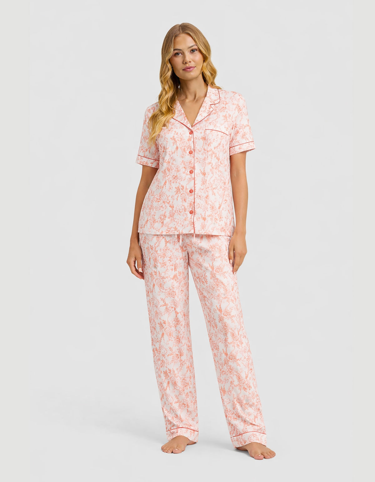 A woman is wearing the Cozy Earth Women’s Bamboo Stretch-Knit Short Sleeve & Pant Pajama Set in Melon Toile, featuring a short-sleeve button-up top and long pants, standing barefoot against a plain light gray background.
