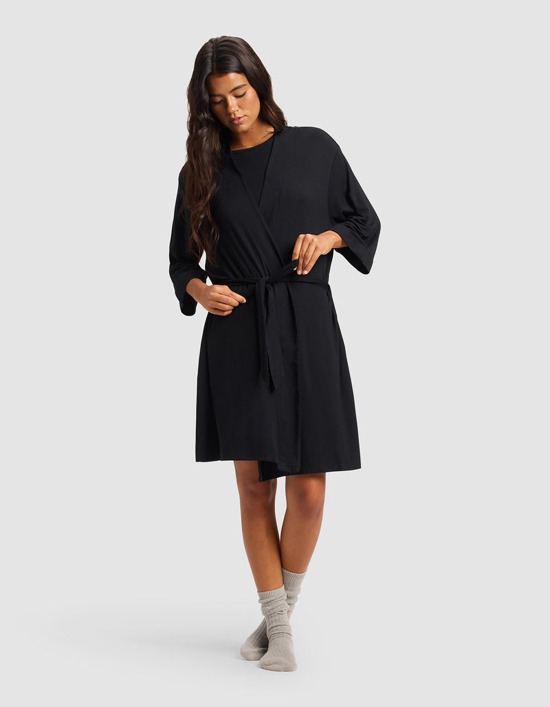 A woman with long dark hair wears the Cozy Earth Women's Bamboo Stretch-Knit Kimono Robe in black, tied at the waist, along with light gray socks. She stands against a plain light gray background, looking down as she gently adjusts the robe's belt.