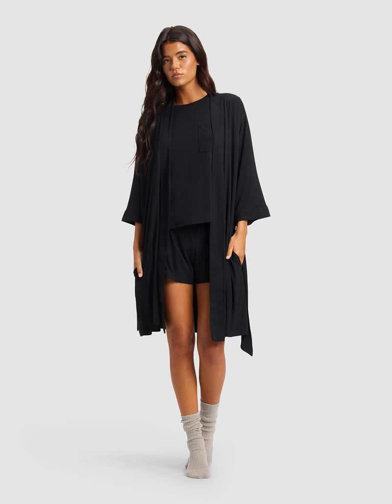 A woman with long dark hair wears the Cozy Earth Women's Bamboo Stretch-Knit Kimono Robe over a black top and shorts, paired with light gray socks, standing against a plain light gray background.