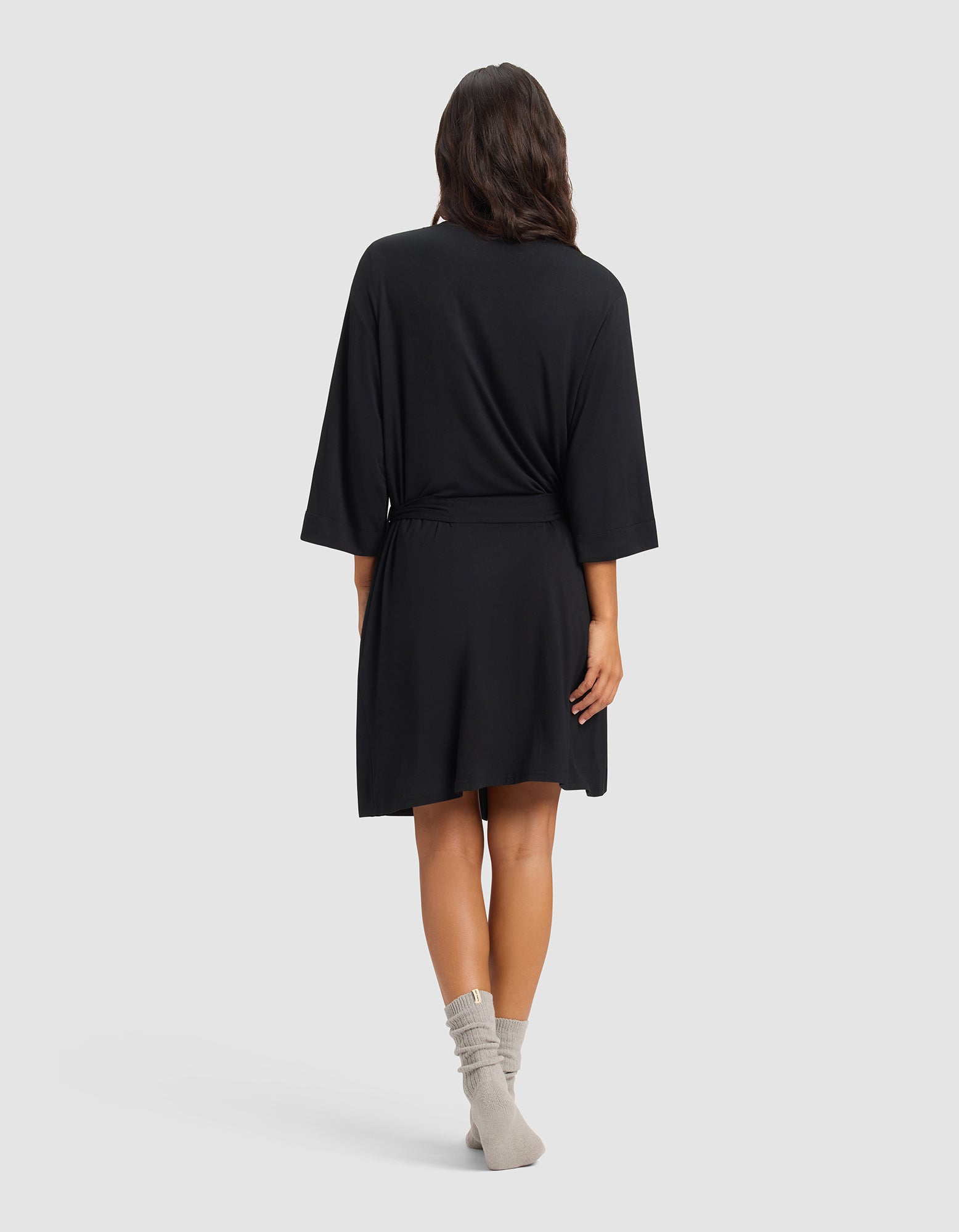 A woman faces away, wearing the Cozy Earth Women's Bamboo Stretch-Knit Kimono Robe in black with a waist tie and gray socks. Her long dark hair drapes over her shoulders against a plain light gray background.