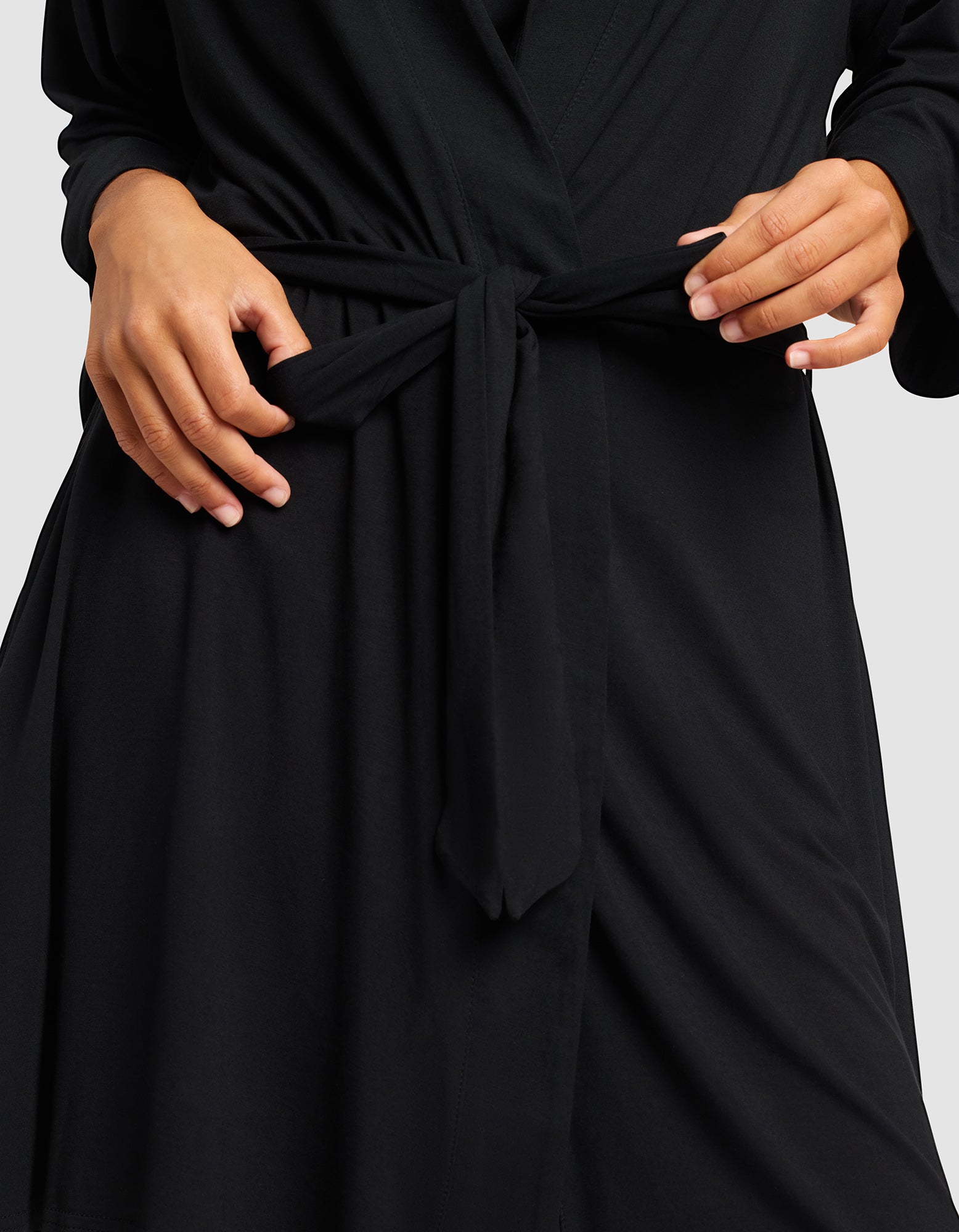 A person adjusts the waist tie of a Cozy Earth Women's Bamboo Stretch-Knit Kimono Robe, showing only their torso and hands against a plain background.