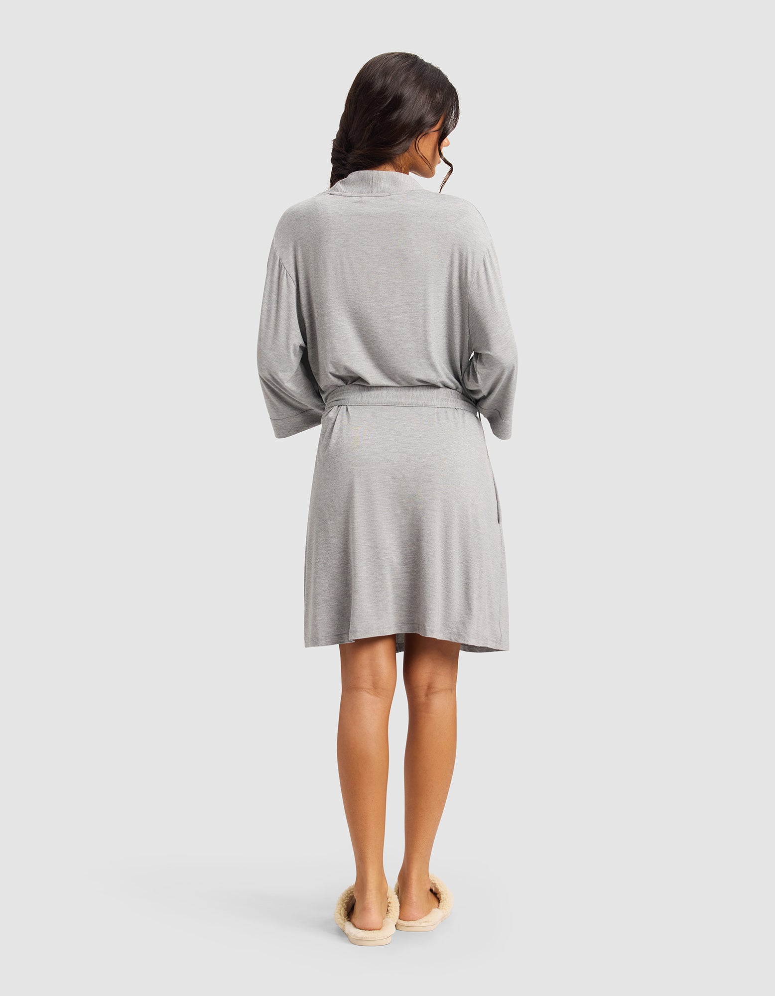 A woman with long dark hair, seen from the back, wears a Cozy Earth Women's Bamboo Stretch-Knit Kimono Robe in light gray with a belt and cream slippers, standing against a plain light background.|Color:Grey