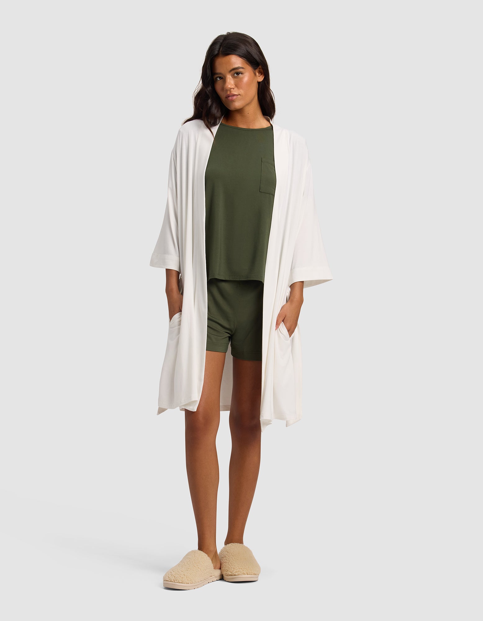 A woman with long dark hair wears the Cozy Earth Women's Bamboo Stretch-Knit Kimono Robe in white over an olive green pajama set with shorts and top, paired with beige slippers, standing against a plain light background.