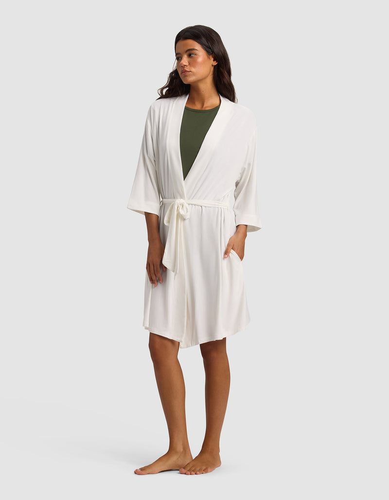 A woman stands barefoot against a light gray background, wearing Cozy Earth's Women's Bamboo Stretch-Knit Kimono Robe in white with a waist tie over an olive green top.
