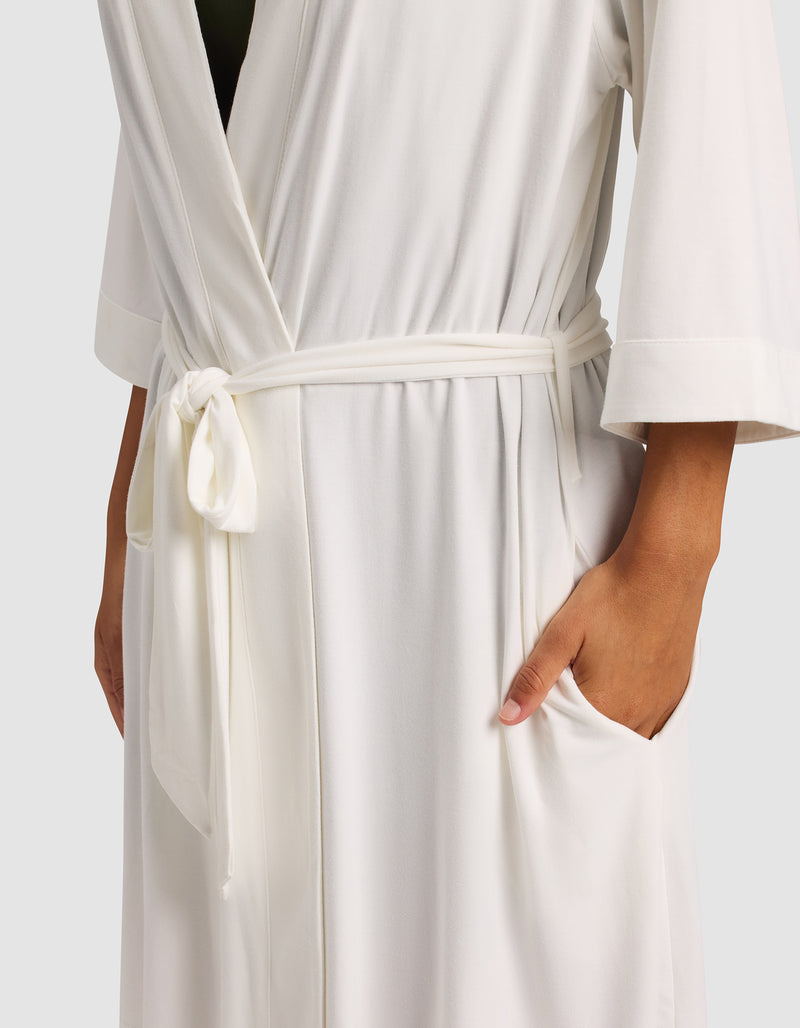 A woman models the Cozy Earth Women's Bamboo Stretch-Knit Kimono Robe—white, tied at the waist with a sash, one hand in a pocket and the other relaxed—against a plain, light background.