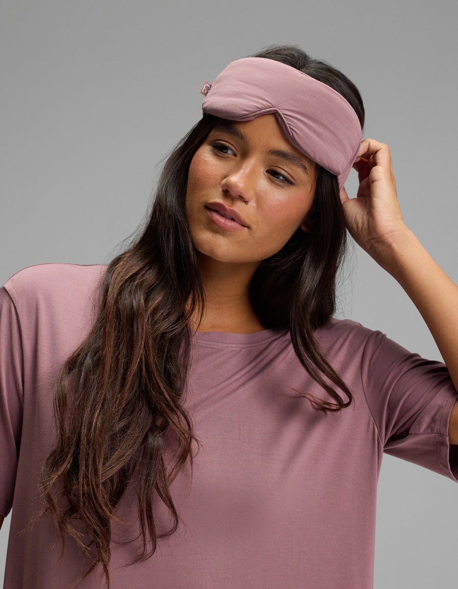 A woman wearing a mauve Bamboo Stretch-Knit Sleep Mask on her forehead and a matching short-sleeved top.