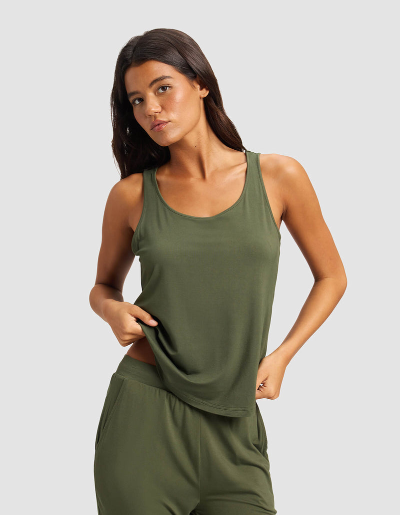 A woman with long dark hair wears the Cozy Earth Women's Bamboo Stretch-Knit Tank & Short Set in olive green, standing against a plain light gray background and slightly lifting the hem of her tank top with one hand.