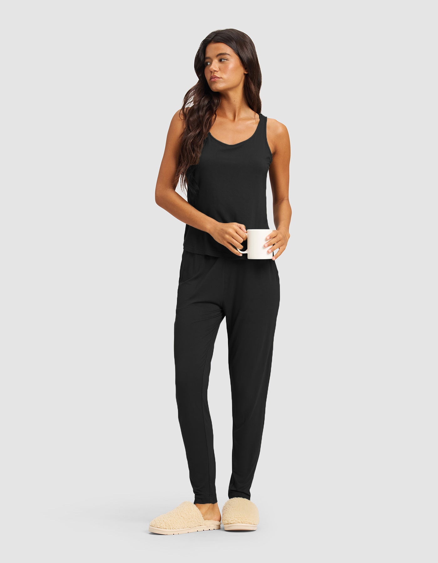 A woman in the Cozy Earth Women's Bamboo Stretch-Knit Tank & Tapered Pant Set, with long brown hair and beige slippers, stands holding a white mug and looking to the side against a plain light gray background.|Color:Black
