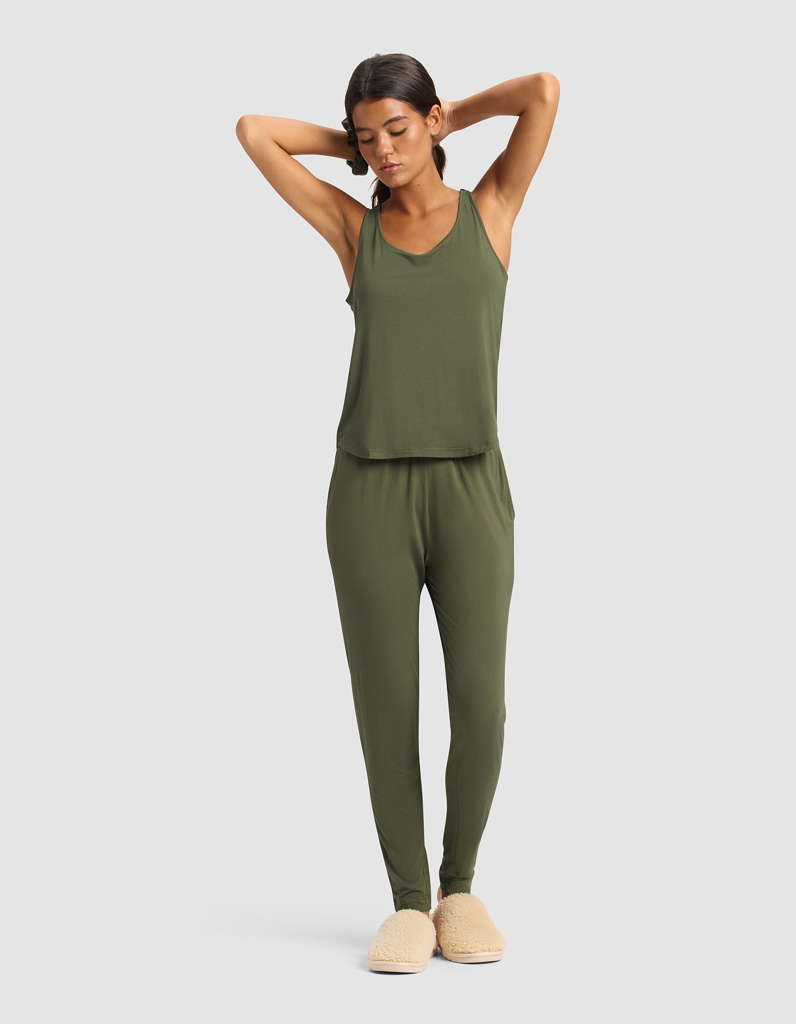 A woman stands with her arms raised, modeling the Cozy Earth Women's Bamboo Stretch-Knit Tank & Tapered Pant Set in olive green and beige slippers against a plain light background.