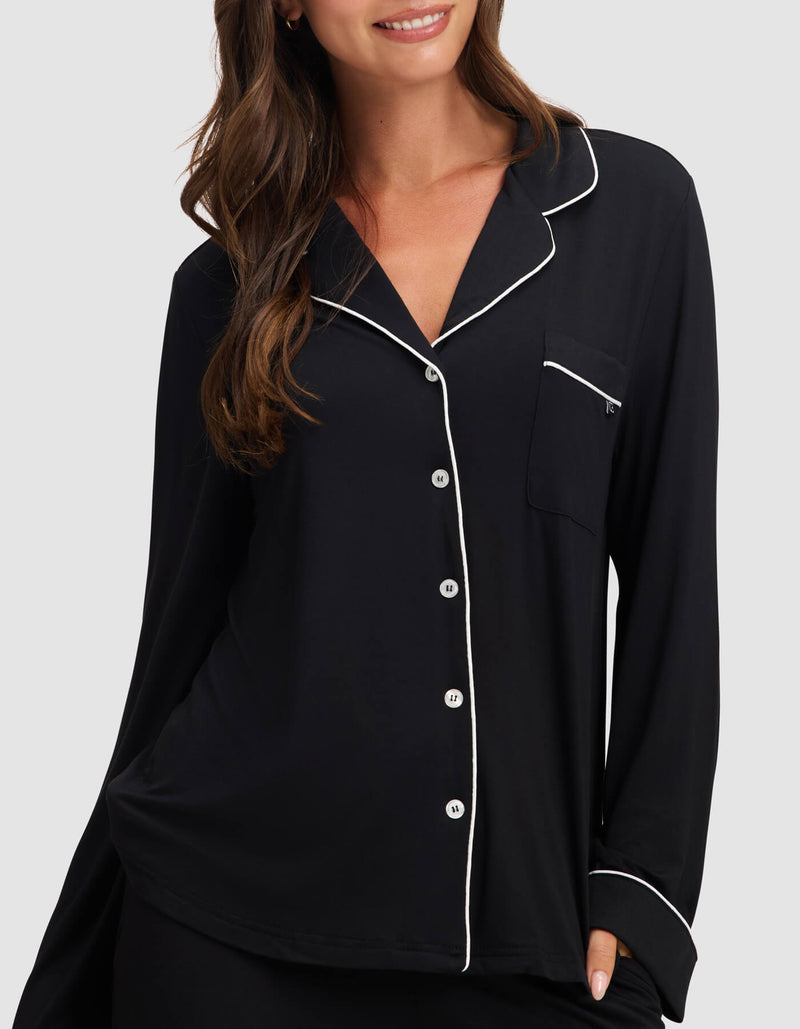 A woman smiles with one hand in her pocket, wearing the Cozy Earth Women's Bamboo Stretch-Knit Long Sleeve Pajama Top in black with white piping, white buttons, and a chest pocket against a plain background.