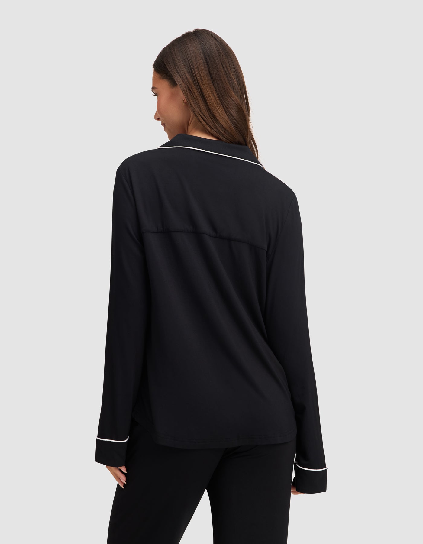 A woman with long brown hair faces away, wearing the HIDE Women's Bamboo Stretch-Knit Long Sleeve Pajama Top in black with white piping, against a plain light gray background. 