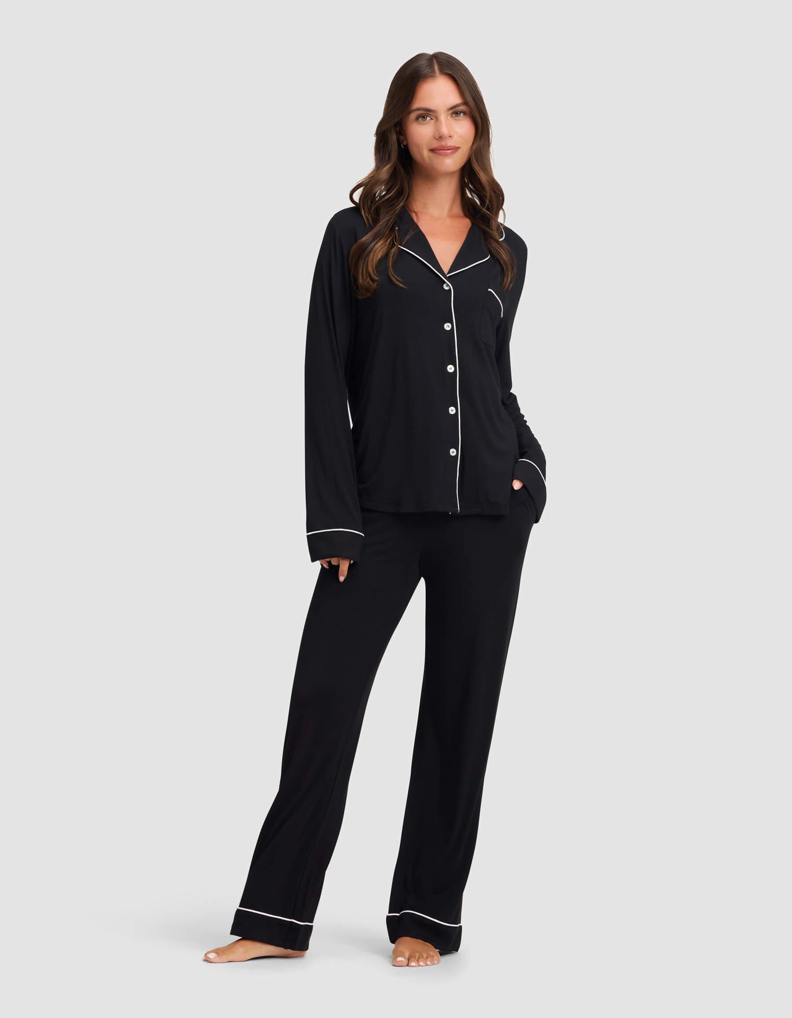 A woman stands barefoot against a light background, smiling slightly with her left hand in her pocket, wearing Cozy Earth's Women's Bamboo Stretch-Knit Long Sleeve Pajama Top in black with white piping and matching pants. 