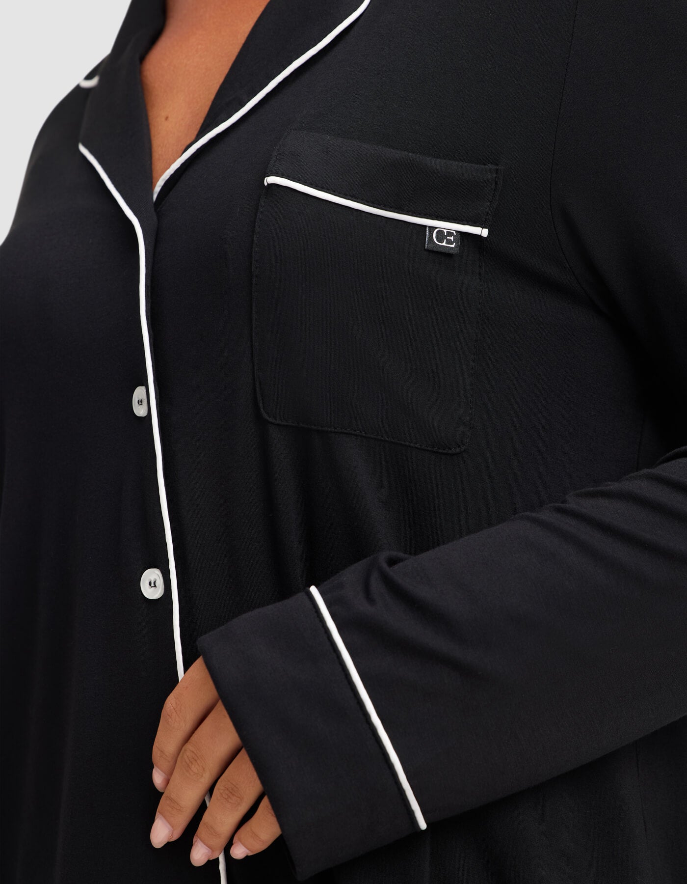 A close-up shows a person wearing the Cozy Earth Women's Bamboo Stretch-Knit Long Sleeve Pajama Top in black, featuring white piping on the collar, pocket, and sleeve, with four white buttons visible and a hand resting near the waist. 