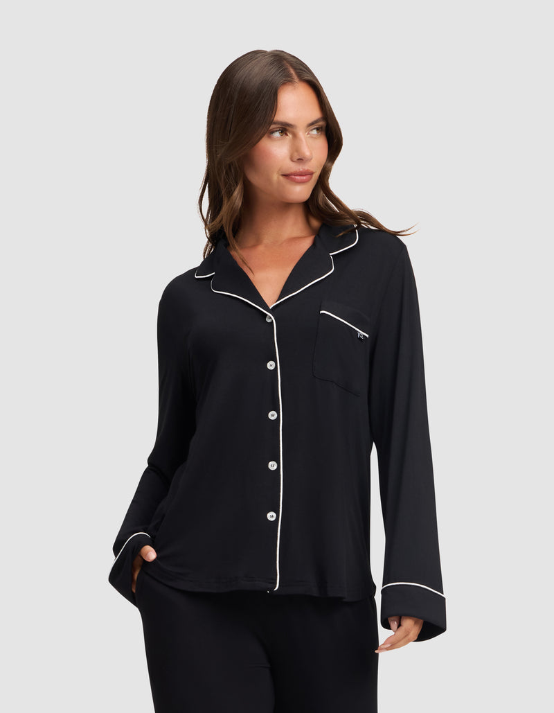 A woman models the HIDE Women's Bamboo Stretch-Knit Long Sleeve Pajama Top in black with white piping on the collar, cuffs, and pocket edges, standing against a light gray background and smiling softly.