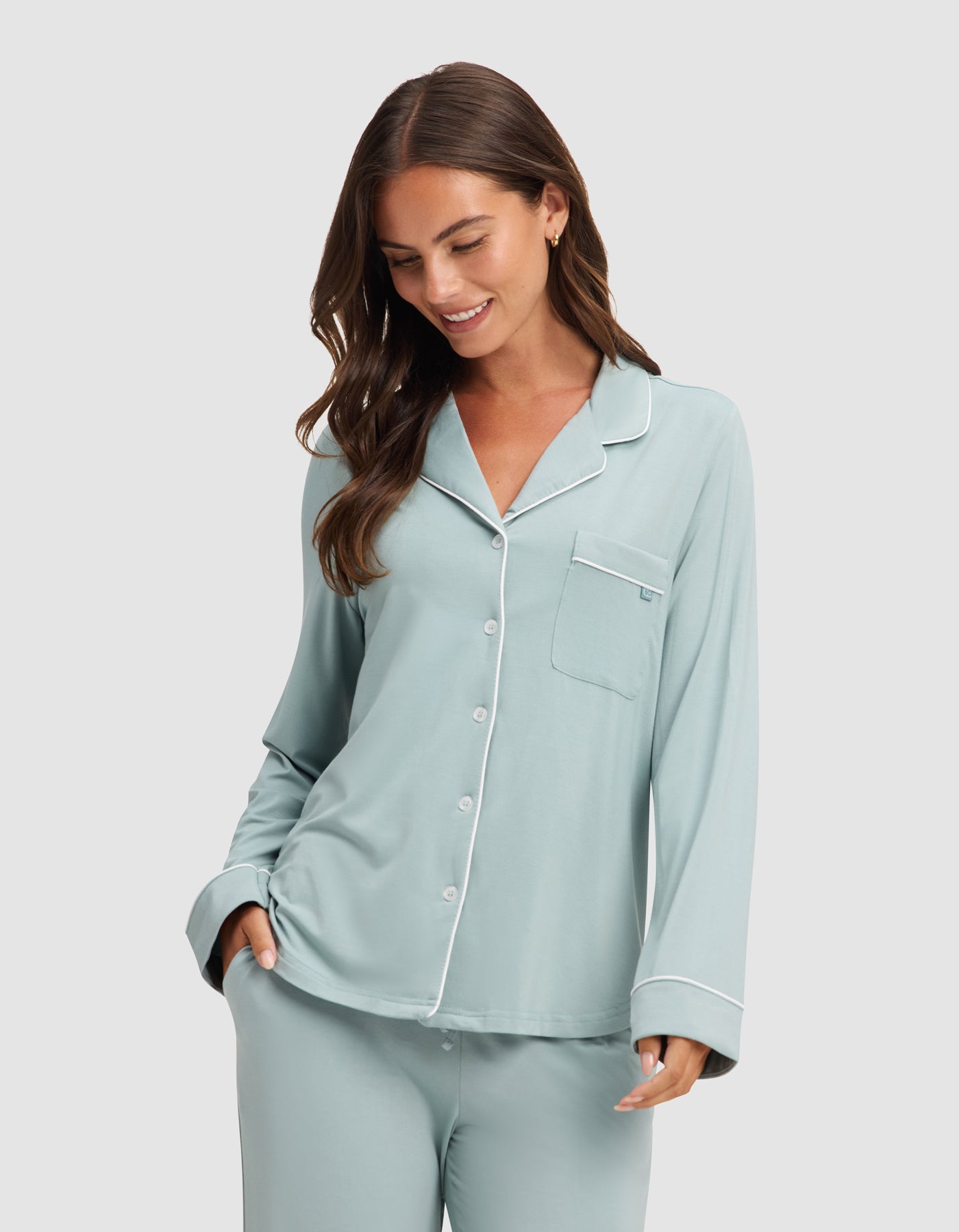 A woman smiles and looks down with one hand in her pocket, wearing HIDE's Women's Bamboo Stretch-Knit Long Sleeve Pajama Top in light blue with white piping and buttons. 