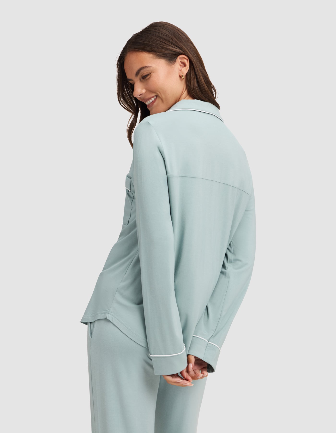A woman wearing the HIDE Women's Bamboo Stretch-Knit Long Sleeve Pajama Top in light blue with white piping smiles over her shoulder against a plain background, her hands relaxed behind her back. 