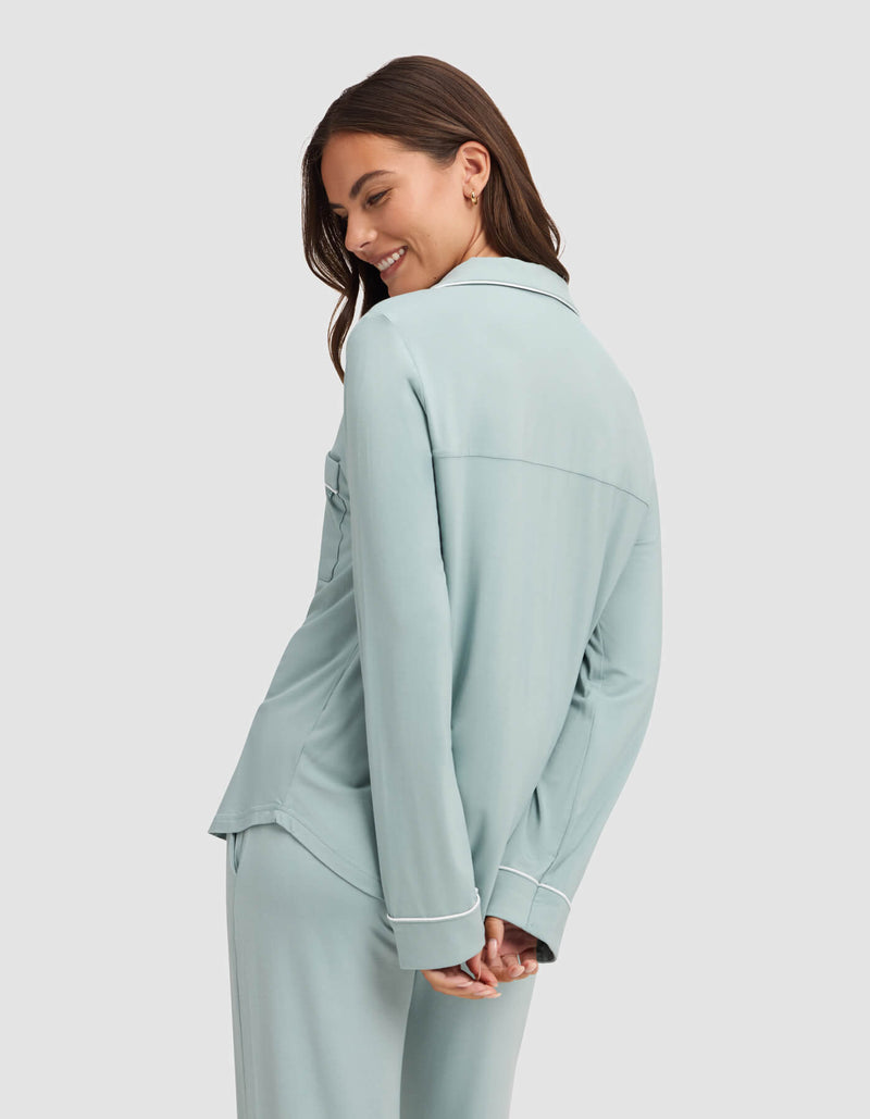 A woman with long brown hair smiles while looking down, wearing the Cozy Earth Women's Bamboo Stretch-Knit Long Sleeve Pajama Top in light blue with white trim, standing against a plain light background.