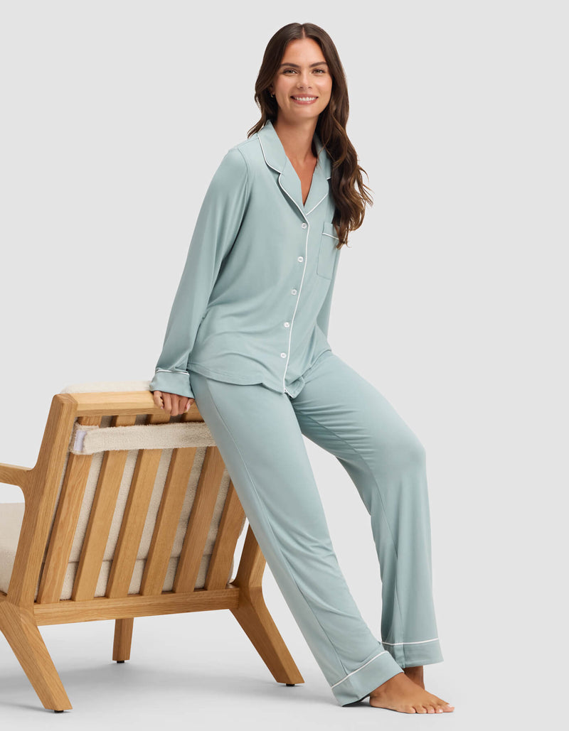 A woman wearing the Cozy Earth Women's Bamboo Stretch-Knit Long Sleeve Pajama Top in light blue smiles while sitting on the armrest of a wooden bench with a cushioned seat against a plain light background.