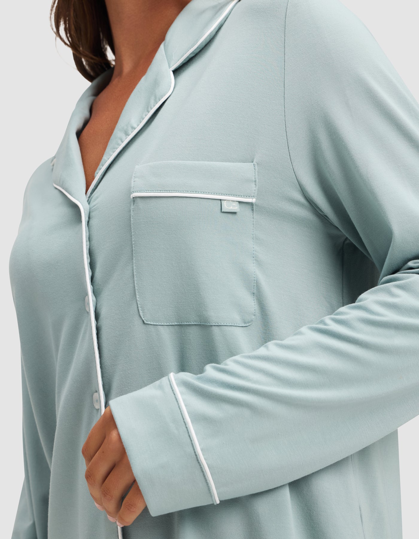 A woman wears the HIDE Women's Bamboo Stretch-Knit Long Sleeve Pajama Top in light blue with white piping, buttons, a front chest pocket, and long sleeves; only her upper body is visible. 