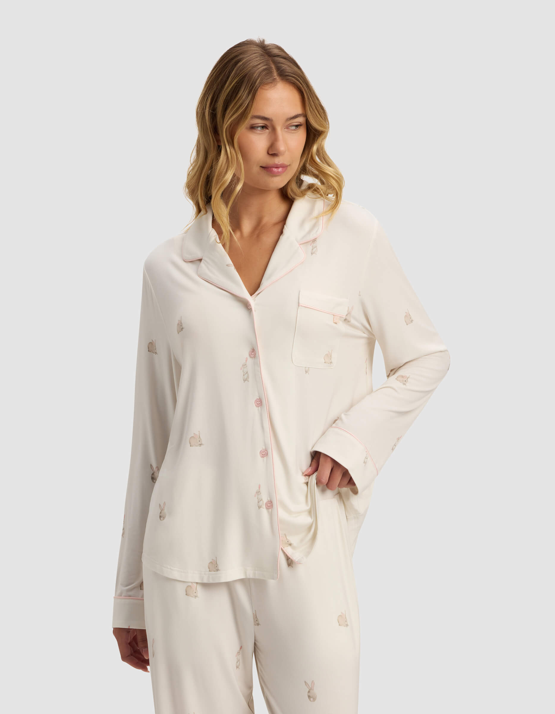 A woman with wavy blonde hair models the Cozy Earth Women's Bamboo Stretch-Knit Long Sleeve Pajama Top in cream with a subtle bunny print, featuring a chest pocket and front buttons. She stands with one hand on her hip, gazing to the side. 