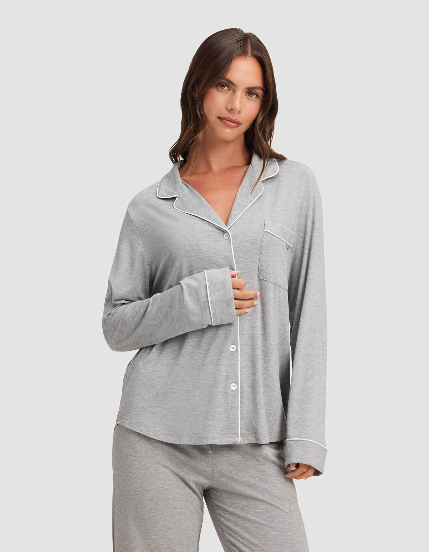 A woman with long brown hair wears the Cozy Earth Women's Bamboo Stretch-Knit Long Sleeve Pajama Top in light gray with white piping, standing against a plain background. She holds the shirt closed with one hand and smiles slightly.