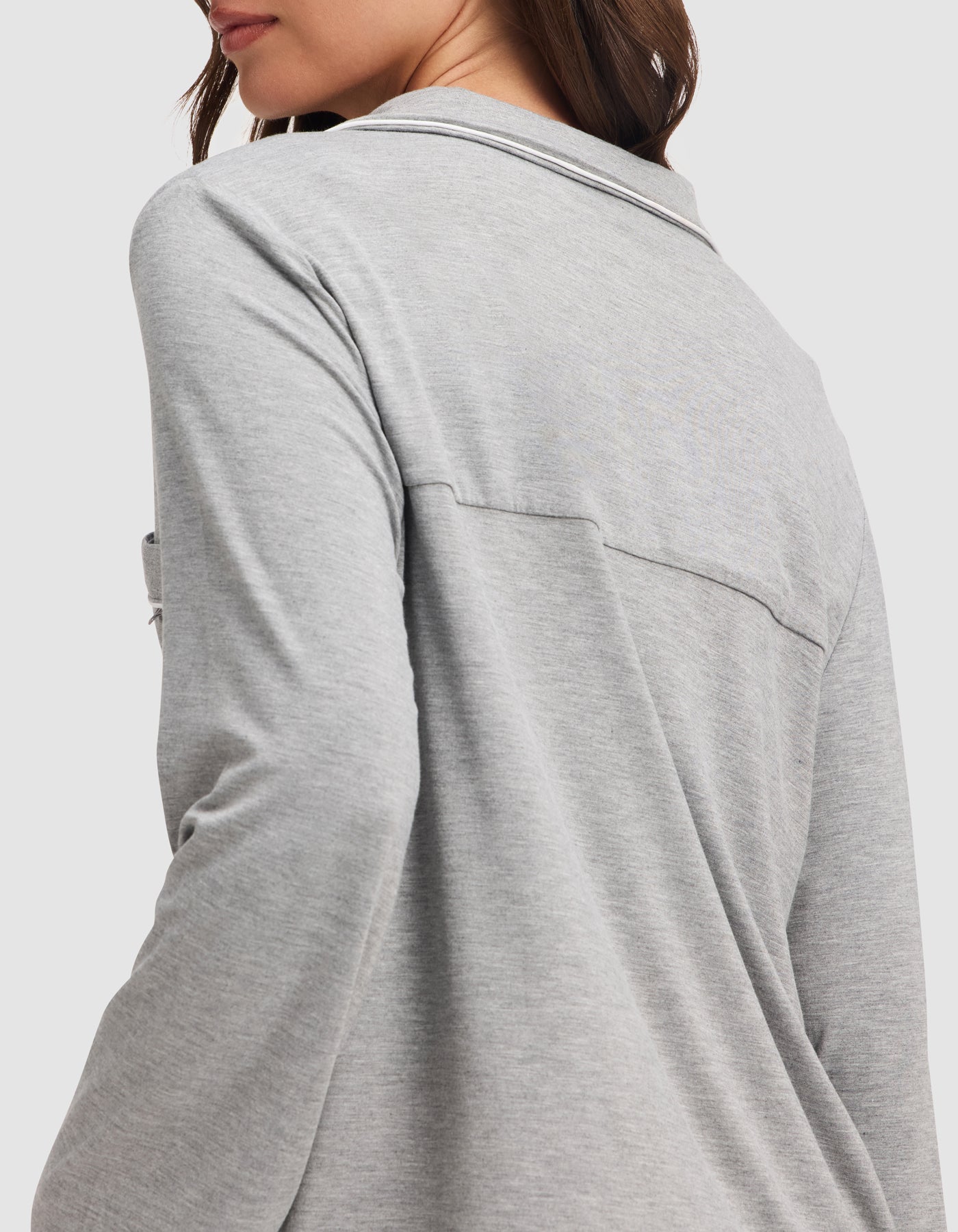 A woman with long brown hair is shown from the side and back, wearing the HIDE Women's Bamboo Stretch-Knit Long Sleeve Pajama Top in light gray. Her face is partially visible against a plain white background. 