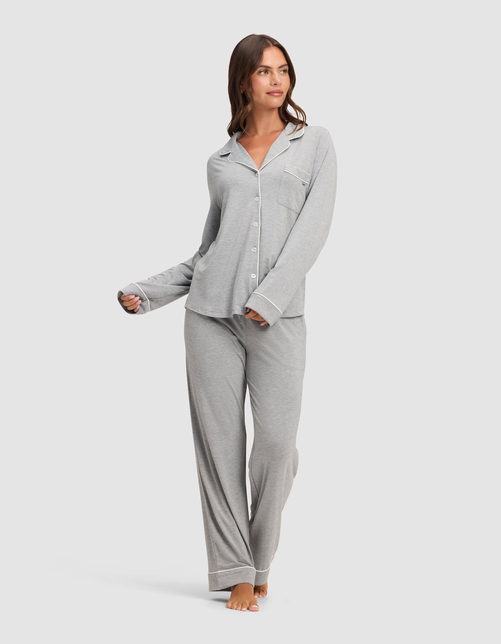A woman stands barefoot against a plain white background, wearing the HIDE Women's Bamboo Stretch-Knit Long Sleeve Pajama Top in light gray, paired with matching loose pants. 