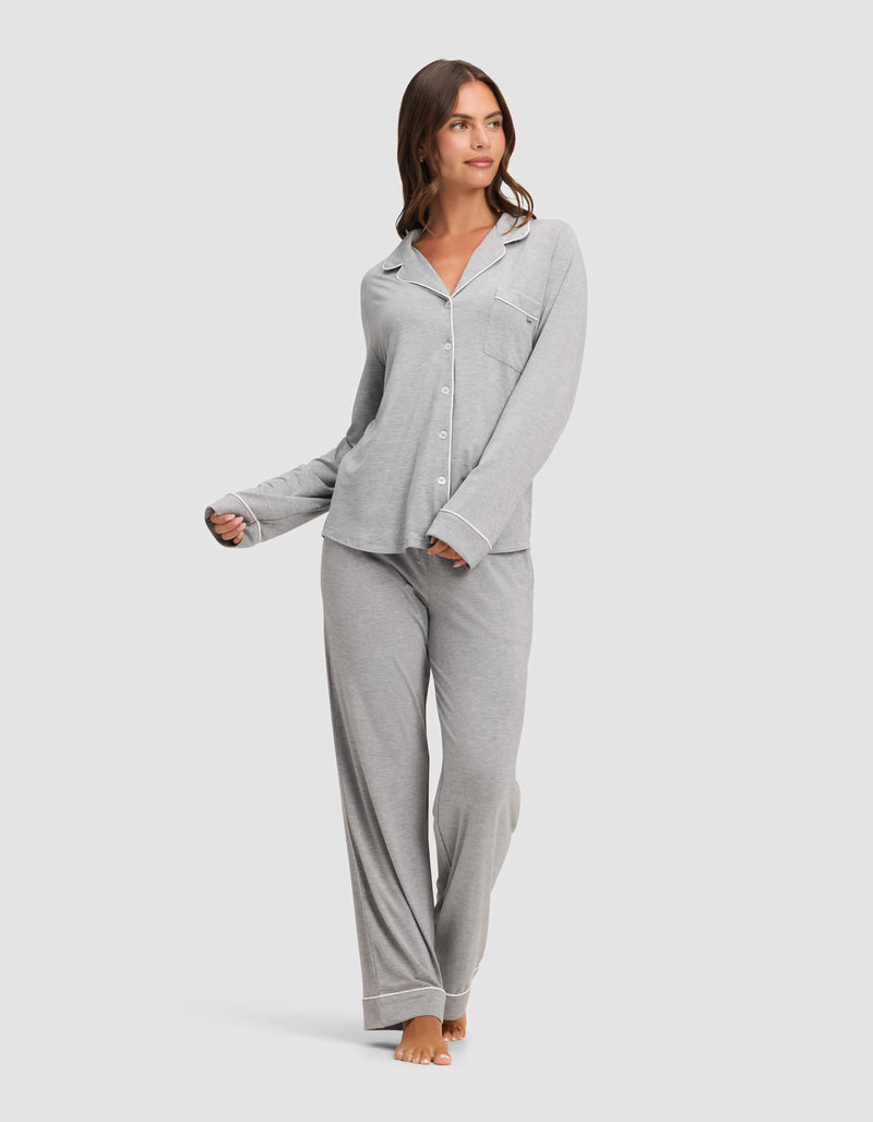 A woman stands barefoot, looking to the side, wearing the Cozy Earth Women's Bamboo Stretch-Knit Long Sleeve Pajama Top in light gray with white trim and a chest pocket, paired with matching loose pants. One hand is slightly raised.