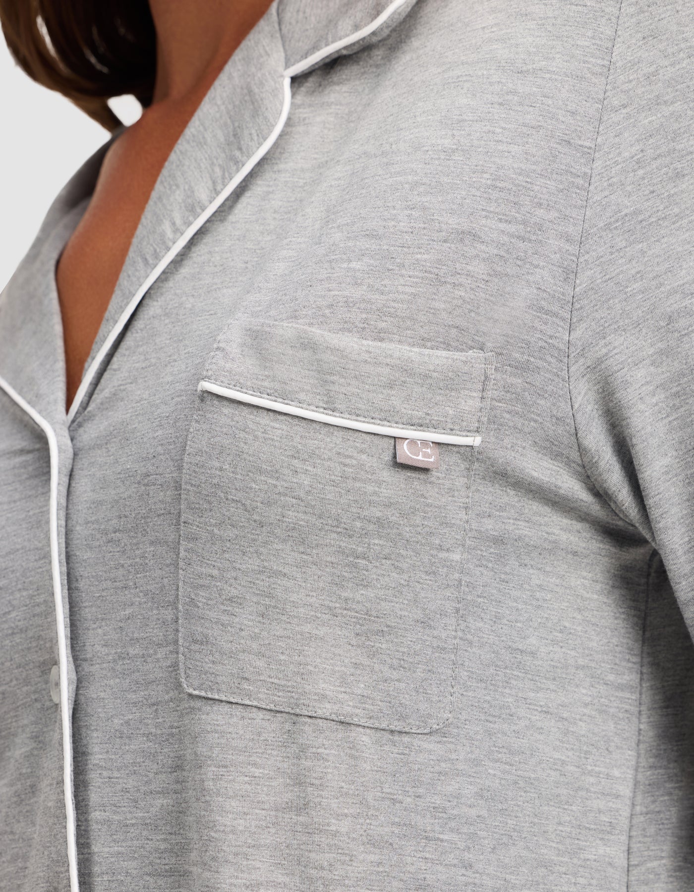 Close-up of a person wearing the HIDE Women's Bamboo Stretch-Knit Long Sleeve Pajama Top in light gray with white piping on the collar, pocket, and trim. The fabric appears soft, and a small tag is visible on the chest pocket's edge. 