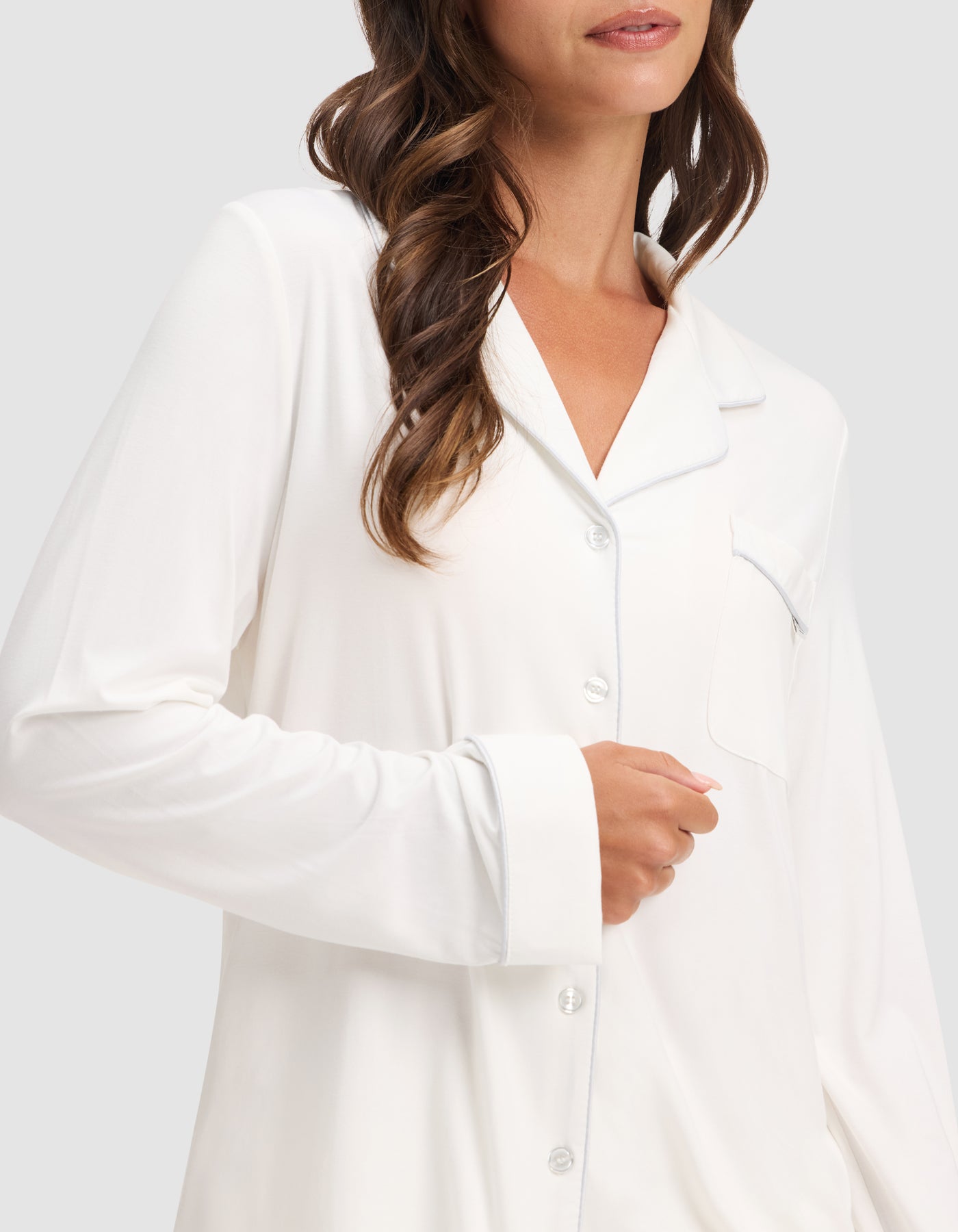 A woman with wavy brown hair wears the HIDE Women's Bamboo Stretch-Knit Long Sleeve Pajama Top, featuring a chest pocket and button-up front. She is shown from shoulders to waist, with her hand resting near the buttons. 