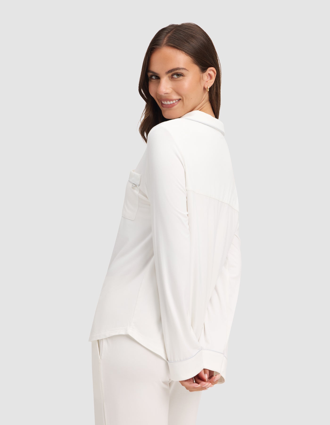 A woman with long brown hair smiles over her shoulder, wearing the HIDE Women's Bamboo Stretch-Knit Long Sleeve Pajama Top in white, paired with matching pants, against a plain light gray background. 