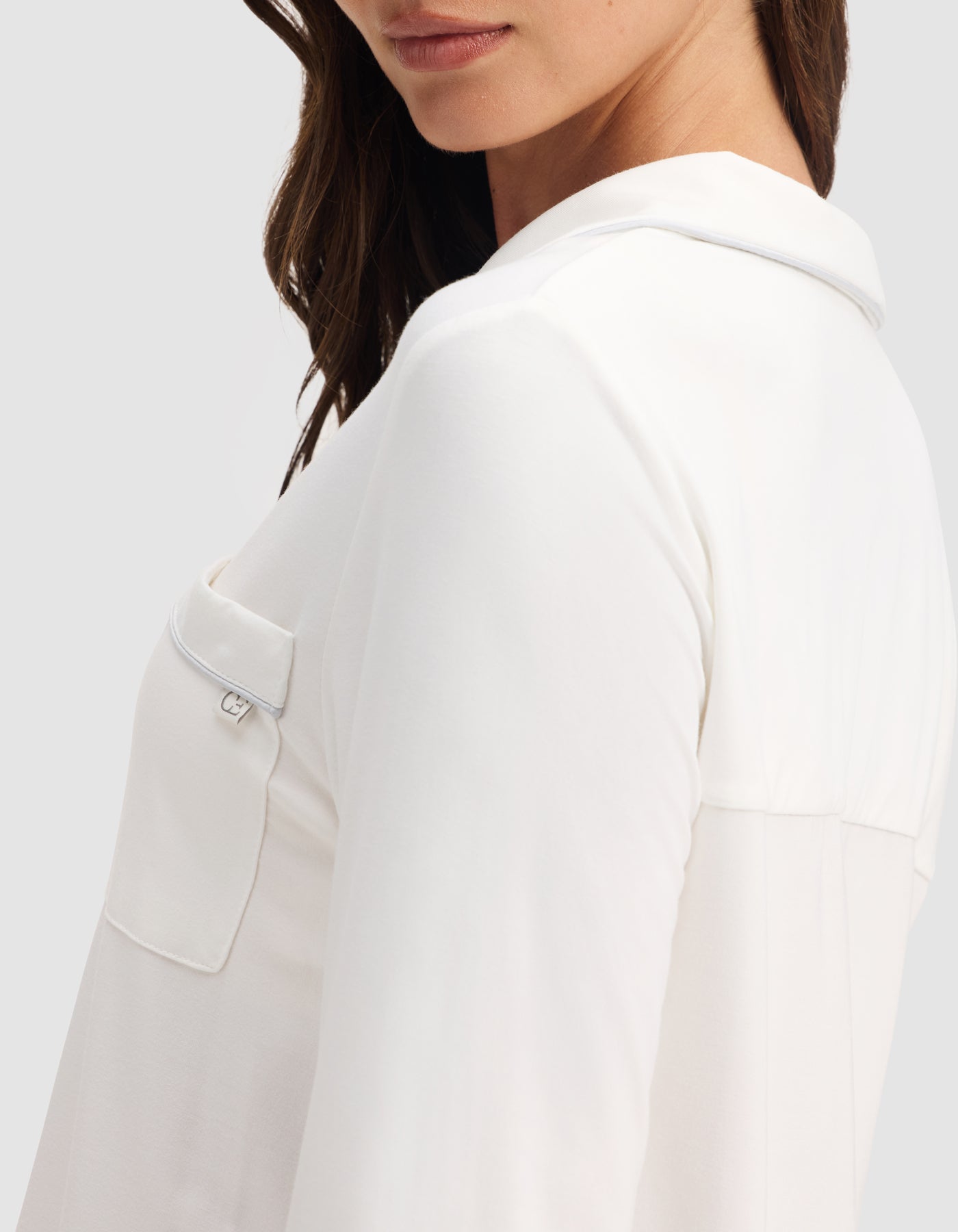 A woman models the HIDE Women's Bamboo Stretch-Knit Long Sleeve Pajama Top in white, featuring a collar and front pocket. The photo is taken from the side at chest height, showing only the lower half of her face and upper body. 