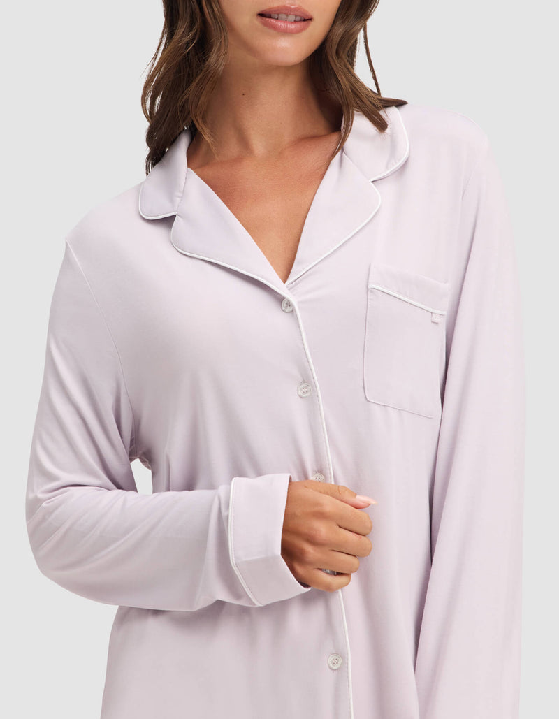 A woman wears the Cozy Earth Women's Bamboo Stretch-Knit Long Sleeve Pajama Top in light lavender with white piping, buttons, and a chest pocket. Her face is partially visible as she holds her right sleeve’s edge.