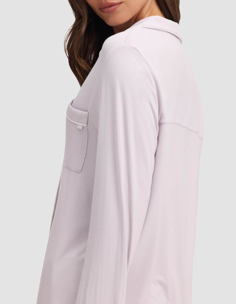 A woman with wavy brown hair models the Cozy Earth Women's Bamboo Stretch-Knit Long Sleeve Pajama Top in light lavender, featuring a chest pocket and button-up front. She is shown from the side against a plain background, her face partly visible.
