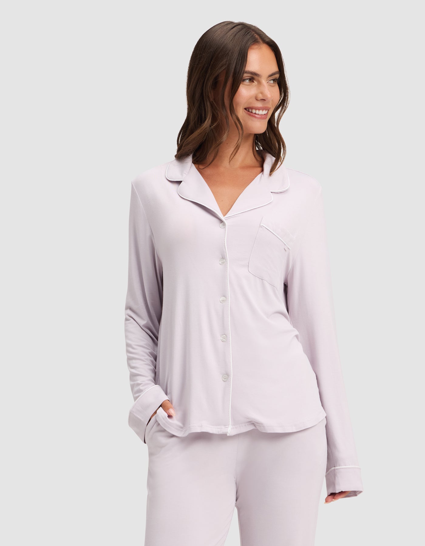 A woman with wavy brown hair smiles and looks to the side, wearing the HIDE Women's Bamboo Stretch-Knit Long Sleeve Pajama Top in light lavender, with her hand in her pocket, standing against a plain light background.  