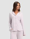 A woman smiles while wearing the Cozy Earth Women's Bamboo Stretch-Knit Long Sleeve Pajama Top in light lavender with a chest pocket, paired with matching pants. She has one hand in her pocket and stands against a plain light gray background.|Color:Lilac