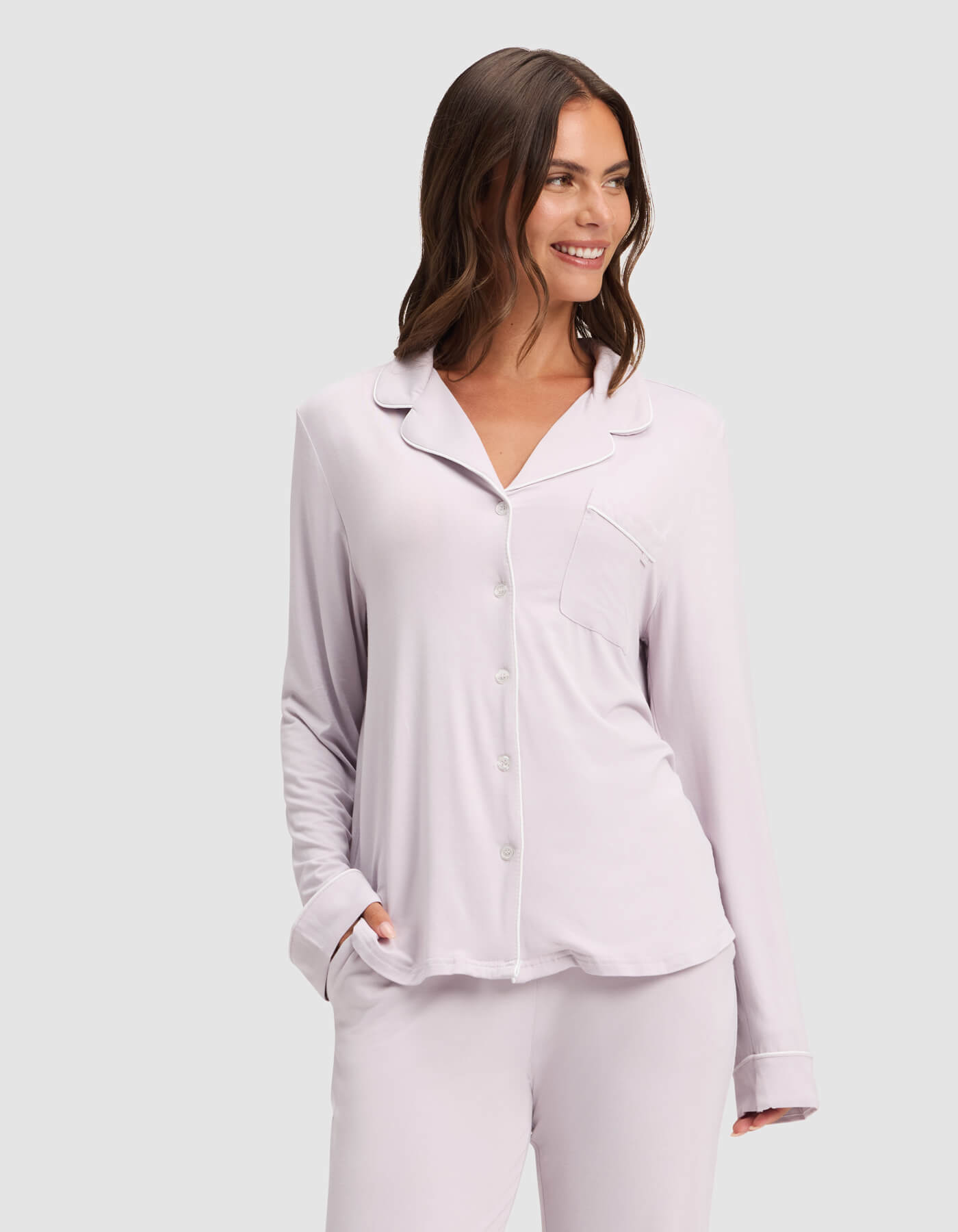 A woman smiles while wearing the Cozy Earth Women's Bamboo Stretch-Knit Long Sleeve Pajama Top in light lavender with a chest pocket, paired with matching pants. She has one hand in her pocket and stands against a plain light gray background.